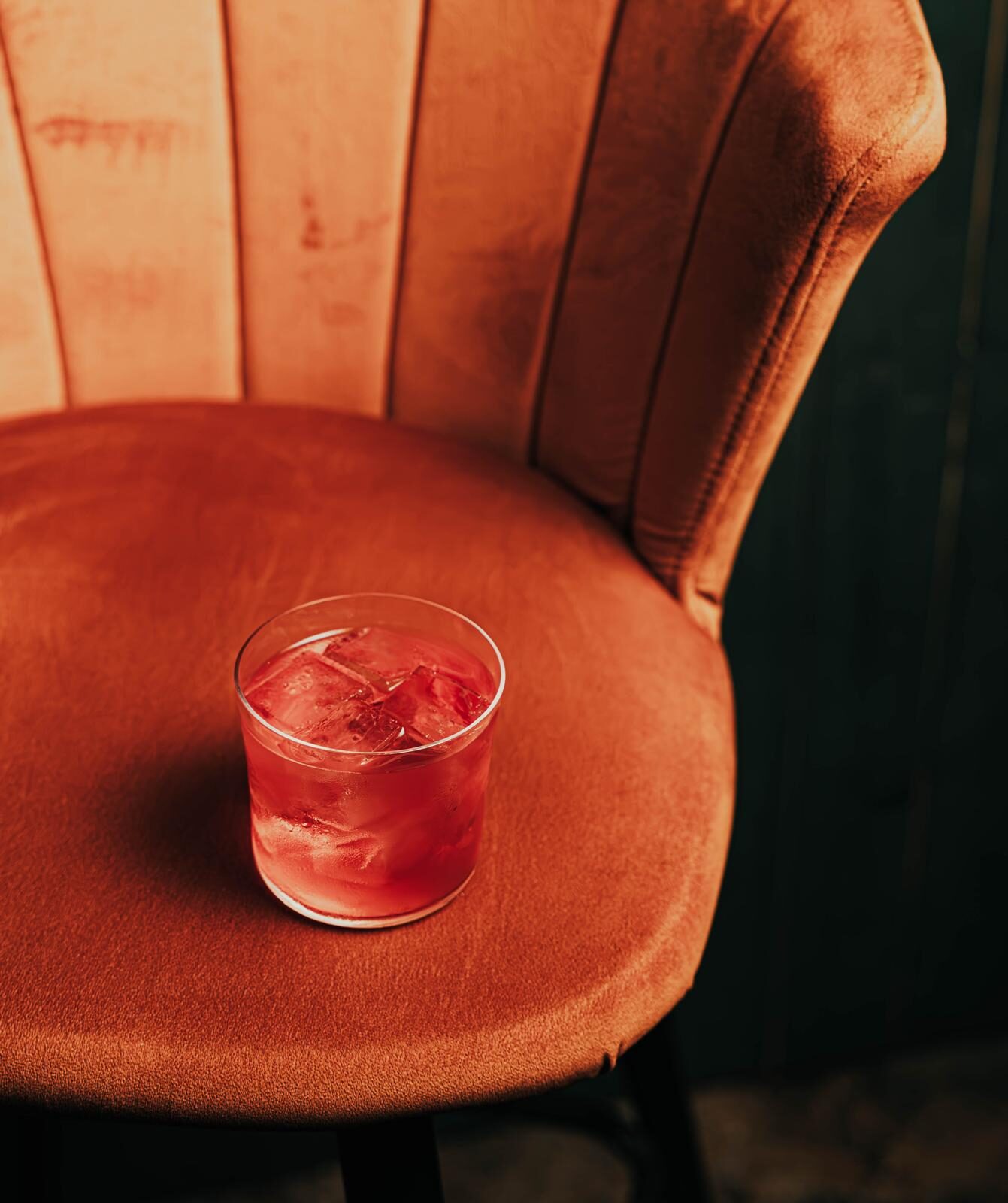 A red cocktail over ice on the burnt orange velvet of an armchair nook